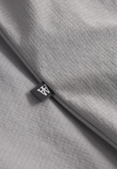 Tissu gris avec une texture côtelée subtile, arborant une étiquette noire avec des lettres blanches. Le design global est minimaliste et épuré.