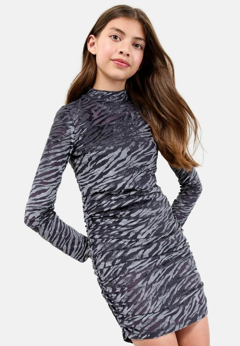Teenagerin mit langen braunen Haaren, die ein eng anliegendes, langärmliges, hochgeschlossenes Kleid mit schwarzem und grauem Tiermuster vor einem weißen Hintergrund trägt.