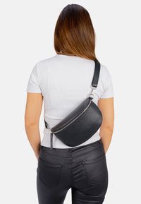 Schwarze Kunstleder-Schultertasche mit geschwungener Form, Reißverschluss und verstellbarem Tragegurt, über dem Rücken über einem weißen, figurbetonten Oberteil getragen.