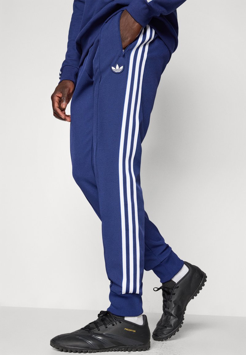 Marineblaue Sweatpants mit weißen Streifen an den Seiten, Reißverschlusstasche und gesticktem Logo, kombiniert mit schwarzen Sportschuhen.