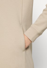 Tissu de pull beige avec une texture lisse, comportant une poche latérale et un poignet ajusté. Détails de couture subtils visibles.