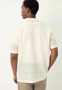 Camisa de manga corta en color crema con un patrón de diamantes texturizado. Presenta un cuello y un ajuste relajado, con un dobladillo trasero ligeramente más largo.