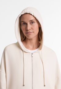 Svijetlosmeđi dukserica na zip s prilagodljivim kapuljačom, slobodnog kroja, srebrnim patentnim zatvaračem i podesivim šniranjem. Glatka tkanina s minimalnim detaljima šavova.