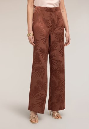 Mujer con pantalones marrones de pierna ancha y cintura alta con patrones de hojas de palma más oscuras, tacones dorados de tiras y una blusa sin mangas de color rosa pálido.