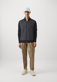 Colete acolchoado preto com fecho de zip, combinado com uma blusa de manga longa escura, calças bege e ténis de cor clara; completado com um boné azul claro.