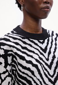 Fekete-fehér zebra mintás pulóver, bordázott fekete nyakkal és textúrált anyaggal, amely elöl cikk-cakk mintát mutat.