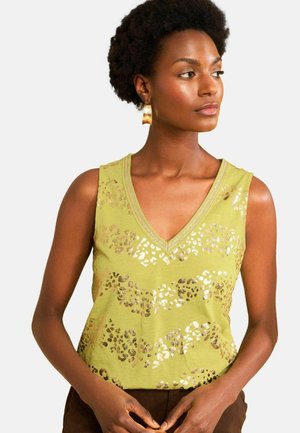 Kobieta z krótkimi kręconymi włosami, nosząca złote kolczyki i zielony bezrękawnik z metalicznym nadrukiem w cętki, patrząca w prawo.