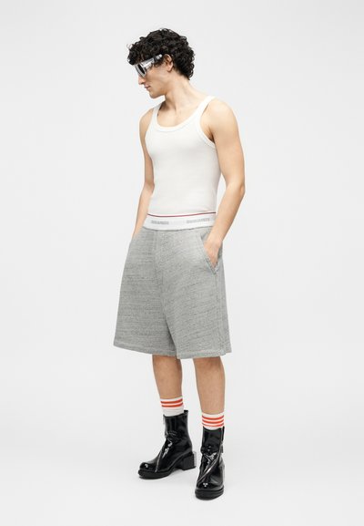 Άνδρας που φοράει λευκό αμάνικο, γκρι μακριά σορτς, ριγέ κάλτσες, μαύρα μποτάκια μέχρι τον αστράγαλο και γυαλιά ηλίου, στέκεται με τα χέρια στις τσέπες κοιτάζοντας αριστερά.