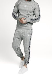 Grå rutigt sweatshirt och matchande joggingbyxor, med svarta sidränder som har varumärket "SikSilk". Bärs med vita sneakers.