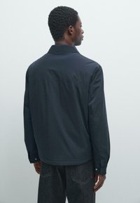 Mand med kort krøllet hår iført en mørkeblå jakke og sorte jeans, står vendt mod en ensartet lys grå baggrund.