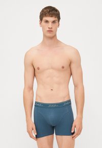 Blaue Baumwoll-Boxershorts mit einem logo-bedruckten Bund; enganliegendes Design mit glatter Textur und Nähten. Modell steht mit einem neutralen Ausdruck.