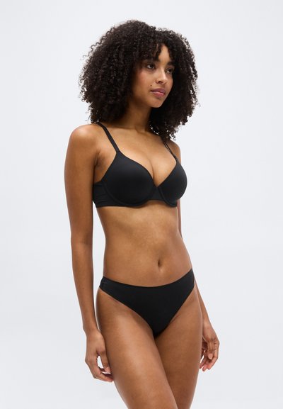Femme aux cheveux bouclés portant un soutien-gorge noir et une culotte noire assortie, debout devant un fond clair uni.