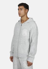 Szara bluza z kapturem na zamek, wykonana z miękkiego materiału, z przednimi kieszeniami, ściągaczami na rękawach oraz wytłoczoną logo na klatce piersiowej. Prosty design.