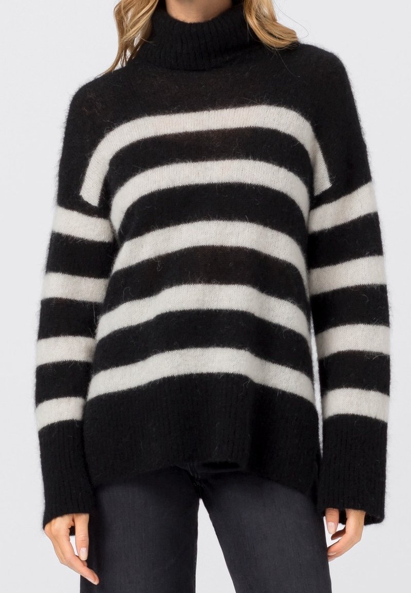 Femme portant un pull à col roulé à rayures floues noir et blanc avec manches longues, associé à un pantalon noir.