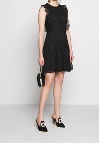 Mustakankaiset mekko, jossa on kukkakuviota, röyhelösoljet ja levenevä helma. Yhdistetty mustiin mokkanahkaisiin muloihin, joissa on korupinnoitteet sekä teksturoitu clutch-laukku.