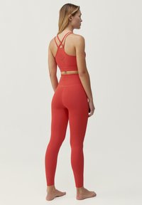 Röd idrottsbehå med korsade axelband och perforerade detaljer, tillsammans med röda högmidjade leggings med subtila mönster och en slät textur.