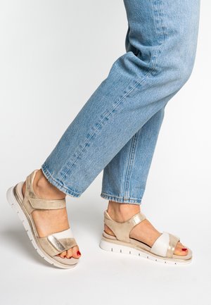 Sandales beige et métalliques à bout ouvert, brides ajustables, accent blanc et semelle blanche rembourrée ; portées avec un jean bleu retroussé.