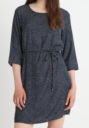 Mujer con un minivestido azul marino oscuro con lunares blancos, mangas tres cuartos y un cinturón fino en la cintura.