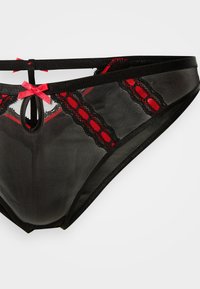 Μαύρο στρινγκ από δαντέλα με δίχτυ, κόκκινες λεπτομέρειες και μικρά φιόγκους. Διαθέτει σχέδιο με ανοίγματα και περίπλοκο μοτίβο δαντέλας κατά μήκος των άκρων.