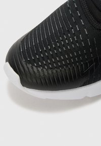 Gros plan sur l'avant d'une chaussure de sport noire avec tissu texturé et caoutchouc, affichant un subtil logo Nike et une semelle blanche.