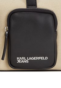 Schwarzlederne Reißverschlusstasche an einer beigefarbenen Canvas-Tasche. Mit silbernem "KARL LAGERFELD JEANS"-Logo. Rundliche Kanten und ein sichtbarer Tragegriff.
