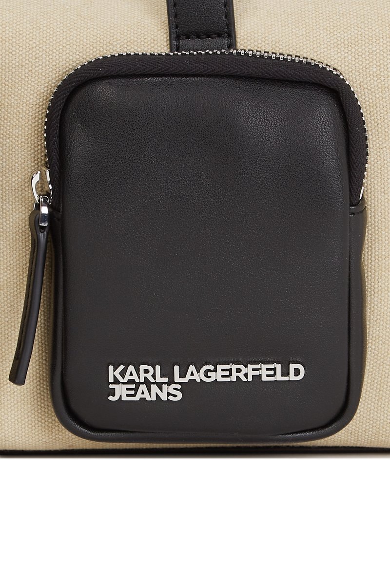 Czarna skórzana kieszeń na zamek błyskawiczny na beżowej torbie z płótna. Posiada srebrne logo "KARL LAGERFELD JEANS". Widoczne zaokrąglone krawędzie i uchwyt na górze.