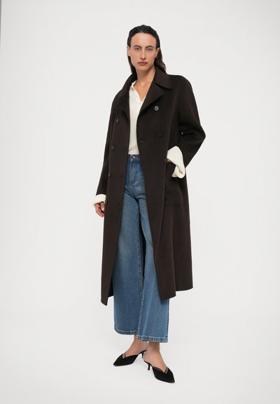 Kvinde iført en lang mørkebrun knapjakke, hvid sweater, vide blå jeans og sorte spidse hæle, stående mod en ensfarvet baggrund.