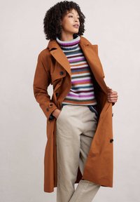 Brązowy płaszcz trench z czarnymi guzikami, noszony otwarty na wielokolorowy sweter w paski. Model ma także na sobie beżowe spodnie z kordurowej tkaniny.