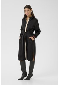Rochie din satin negru cu mâneci lungi, un plastron cu butoane bej contrastant, cordon în talie, fante laterale și purtată cu cizme negre până la genunchi.