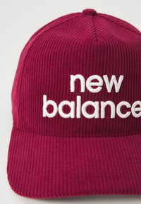 Bordó kordbársony sapka fehér hímzett "new balance" felirattal. Gömbölyített korona és ívelt karima jellemzi. A textúrázott szövet részletei láthatóak.