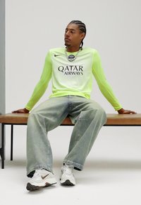 Neona zaļš garām piedurknēm krekls ar melnu logo, valkāts kopā ar gaiši ziliem brīva piegriezuma džinsiem un baltiem sporta apaviem. Persona sēž uz koka sola.