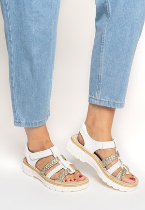 Füße in weißen und goldfarbenen Sandalen mit geflochtenen Riemen und Klettverschlüssen, kombiniert mit hellblauen, hochgekrempelten Jeans.