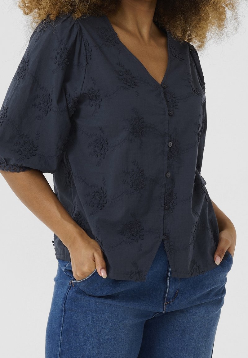 Femme portant un chemisier bleu marine boutonné avec broderie florale et manches bouffantes, associé à un jean en denim bleu, mains dans les poches.
