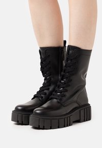 Botas de tobillo de cuero negro con una suela de goma gruesa, que cuentan con cordones y una cremallera lateral para facilitar su uso. Textura suave y punta redonda.