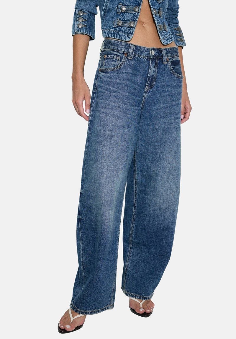 Femme portant un jean bleu ample, une veste en jean courte avec des boutons, et des sandales tongs noires avec des brides blanches.