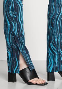 Calças com um padrão de ondas azul e preto, apresentando fendas laterais. Combinadas com uma sandália preta de salto bloco com design de ponta aberta.