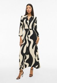 Dolga črna in smetanasta obleka z abstraktnim valovnim vzorcem, gumbi spredaj in tekočim dizajnom. Dopolnjena z odprtimi črnimi petkami.