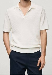 Polo blanc en maille avec manches courtes, col en V et base côtelée. Associé à un pantalon bleu marine foncé.