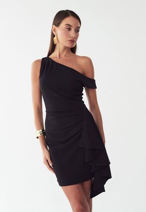 Mujer con un vestido negro de un solo hombro con detalle de volantes superpuestos en el lateral, pendientes dorados y una pulsera dorada, de pie frente a un fondo liso.