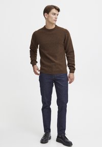 Brauner Strickpullover mit rundem Ausschnitt, kombiniert mit dunkelblauen Jeans und schwarzen Schnürschuhen. Die Textur wirkt weich und gemütlich.