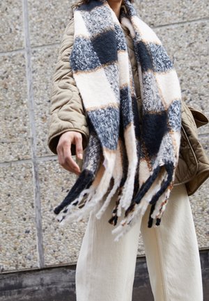 Personne portant une veste matelassée beige et un pantalon crème, tenant une grande écharpe à carreaux noirs, blancs et marron clair avec des franges aux extrémités.