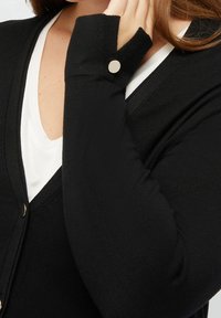 Černý kardigan s dlouhými rukávy, měkká tkanina, design s výstřihem do V a knoflíkovým akcentem na rukávu. Nositelný přes bílé tričko.