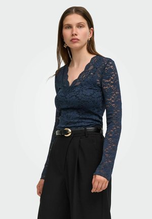 Femme portant un haut en dentelle bleu marine à manches longues avec un col festonné, un pantalon noir taille haute et une ceinture noire avec une boucle dorée.