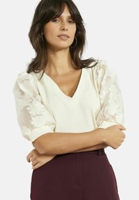 Kreemjas V-kaelusega särk tekstuurse kangaga, millel on läbipaistvad lillemustriga padjavarukad ja lõdvestunud lõige, sobitatud tumepunaste pükstega.