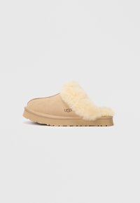 Tan semskinnslipper med en myk, kremfarget pelskant og en flat, teksturert gummisåle. Har et "UGG"-logo på siden.