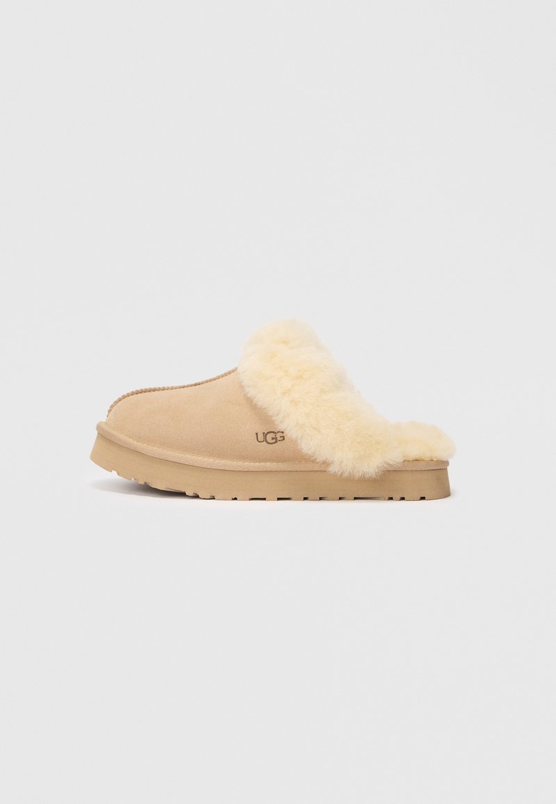 Tan semskinnslipper med en myk, kremfarget pelskant og en flat, teksturert gummisåle. Har et "UGG"-logo på siden.