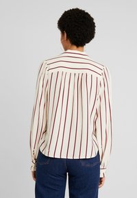 Blusa crema con rayas verticales en color burdeos, mangas largas abullonadas, puños abotonados y detalle de pliegues en la espalda. Combinada con jeans azules.