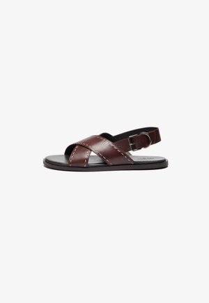 Brown leather sandals with cross straps, a buckle on the ankle, and a flat black sole. Stitched detailing along the straps.