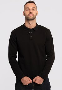 Svart långärmad polo med krage, med tre knappar fram och textilstruktur. Bärs över mörka denimjeans.