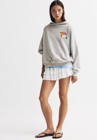 Sweatshirt gris oversize avec un graphique de fruits colorés, mini jupe blanche à volants et sandales noires. Tissu doux, design décontracté.